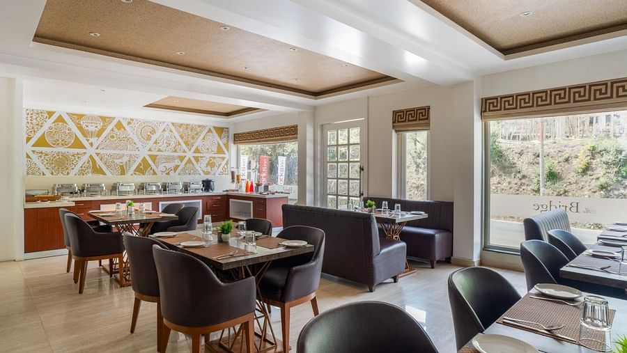 The Bridge restaurant at Clarks Inn, Near Kainchi Dham features set tables, a buffet area, booth seating, large windows, and decorative wall and ceiling details.