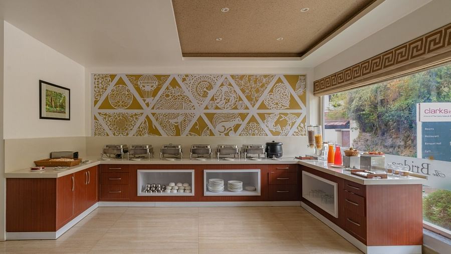 The breakfast buffet area at The Bridge restaurant, Clarks Inn, Near Kainchi Dham features a selection of covered dishes, drinks dispensers, plates, and decorative wall.