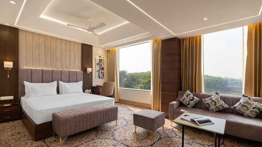 A luxurious hotel suite with a king-size bed, an elegant brown upholstered headboard, and a plush carpet. A large window provides a view of greenery outside. A grey sofa with decorative pillows, a coffee table, and a small footstool complete the seating ar