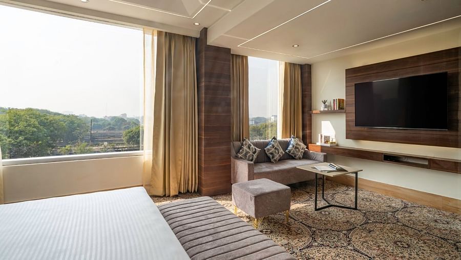 A hotel suite with a king-size bed facing a large window that offers a scenic view of lush greenery. The room features a grey sofa, a wall-mounted television, and a wooden console with decorative shelving. The carpet has an intricate design, and the ceilin