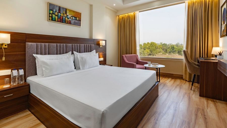 Image 1: A modern hotel room featuring a large double bed with white linens, a wooden headboard, and two bedside tables with lamps. A window with beige curtains lets in natural light, and a seating area with a pink armchair and a small round table is near 