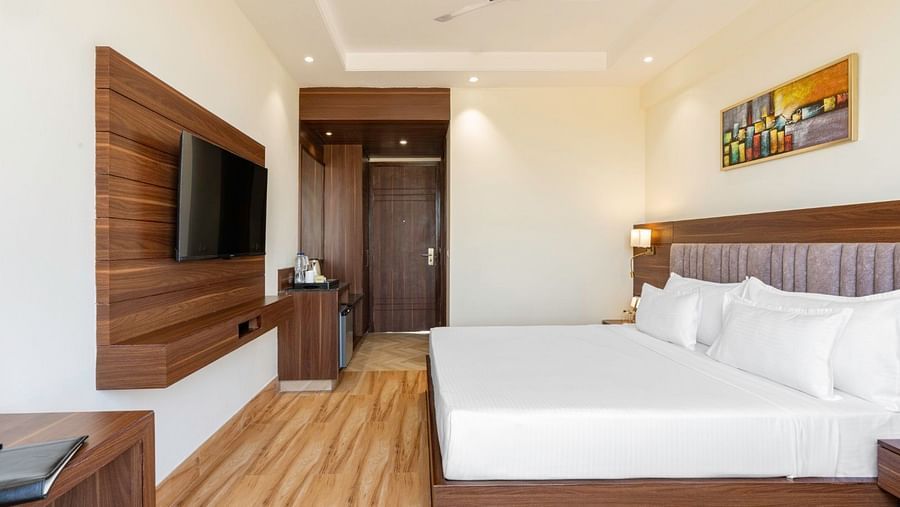 A side view of a modern hotel room with a large double bed covered in white linens, a wooden headboard, and a bedside lamp. A wooden panel on the opposite wall holds a flat-screen television. The entrance door is visible in the background, along with a war