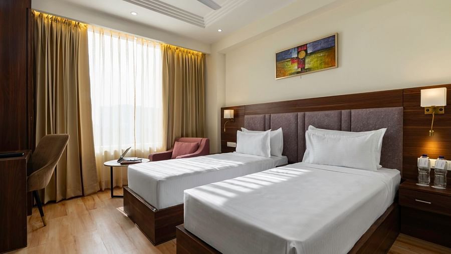 A modern hotel room featuring two single beds with white linens, wooden flooring, and a large window with beige curtains. The room is furnished with a desk, chair, and a seating area with two pink armchairs and a round table. A colourful abstract painting 
