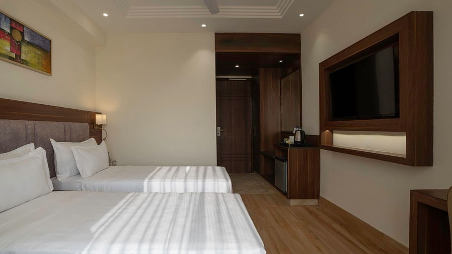A hotel room with two single beds, neatly arranged with striped white bedspreads. A wall-mounted flat-screen television is opposite the beds, framed with wooden paneling. A minibar, kettle, and wardrobe are visible near the entrance. The walls are painted 