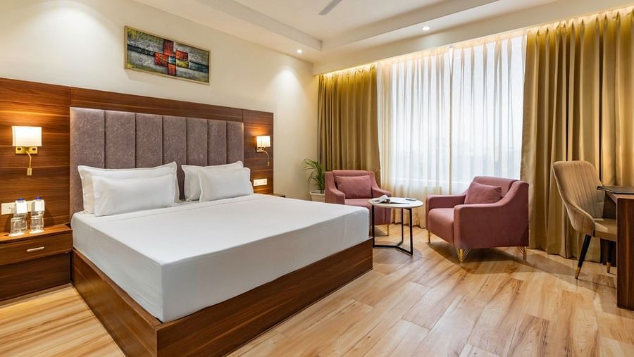 A contemporary hotel room with a large bed, wooden flooring, and a seating area with two pink armchairs and a small round table. A wooden headboard with upholstered panels is illuminated by bedside lamps. A colourful painting adds vibrance to the neutral-t