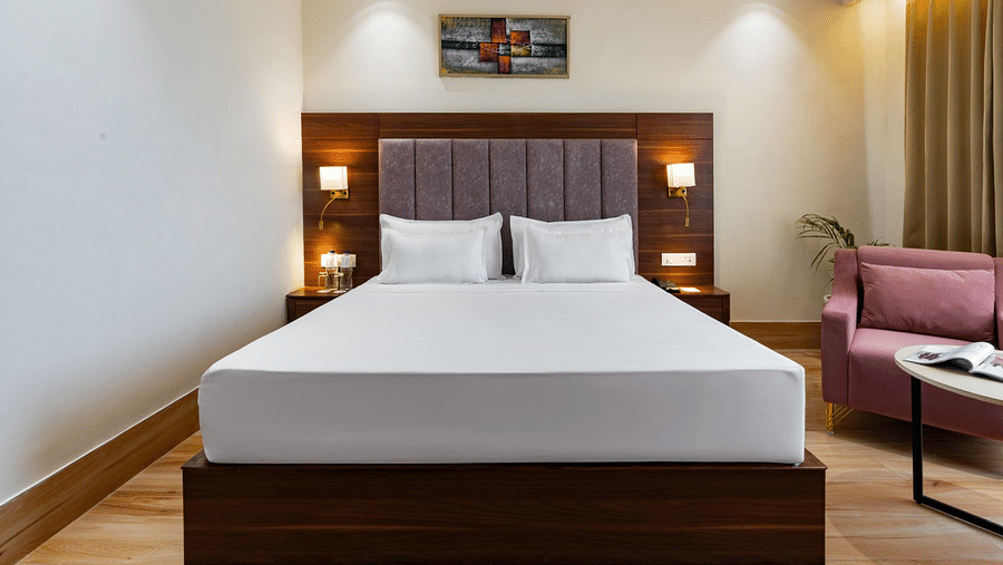 A well-lit hotel room featuring a large bed with a brown upholstered headboard, neatly arranged with white linens. Two bedside lamps cast a warm glow on the wooden paneling. The seating area includes a pink armchair, a round coffee table, and a green potte