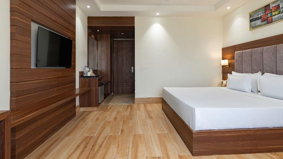 A modern hotel room with a king-size bed, a wooden headboard, and minimalist decor. The room has a large wall-mounted television, a wooden console, and a workspace with a chair. The entrance area leads to a wardrobe and a minibar setup. Soft recessed light
