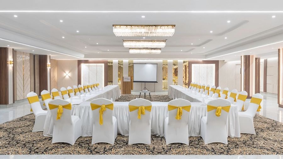 A conference room with white-covered chairs tied with yellow bows, a long U-shaped table, and a projector screen at the front – Clarks Inn Suites, Near Taj Mahal.