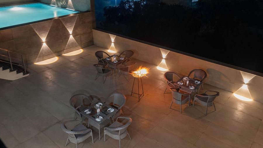 A rooftop dining area at night with neatly arranged tables, rattan chairs, a lit fire pit, and wall-mounted lights – Clarks Inn Suites, Near Taj Mahal.