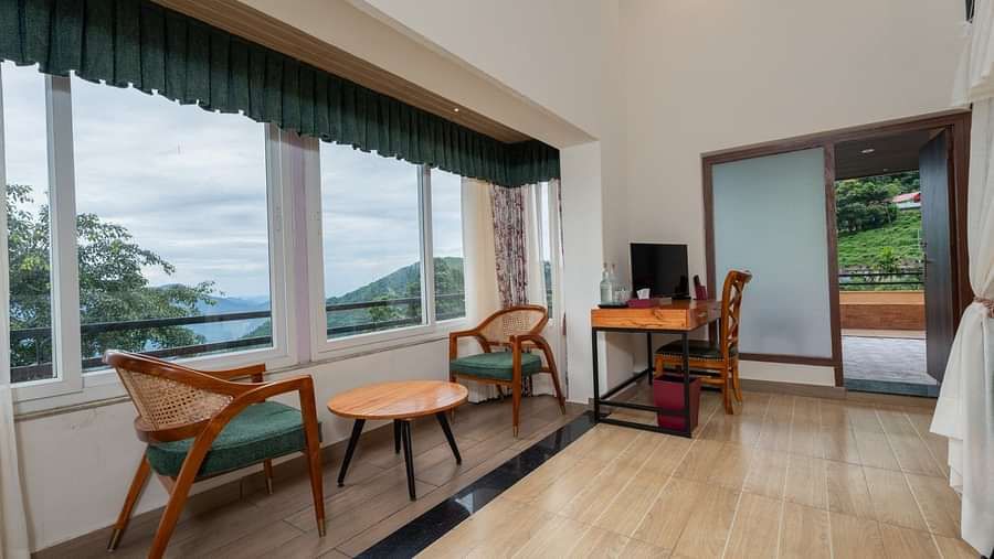 Large windows with nature views, comfortable seating, and a wooden table complement the spacious bed with side curtains and wooden furnishings