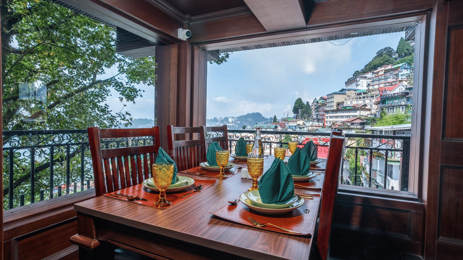 Dining table set with tableware beside large windows looking towards hillside buildings at The West Gate Posada By Summit, Darjeeling.