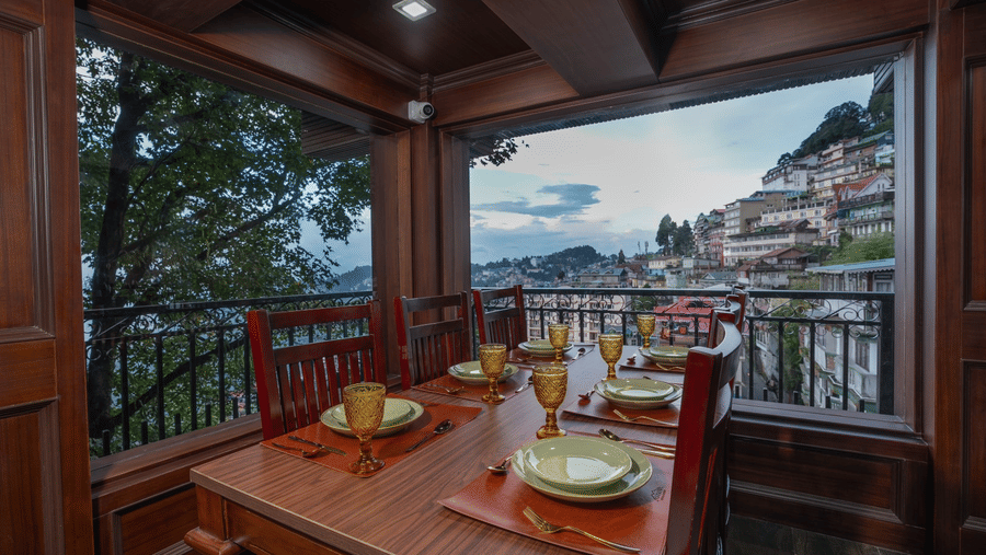 Dining table set with tableware beside large windows looking towards hillside buildings at The West Gate Posada By Summit, Darjeeling.