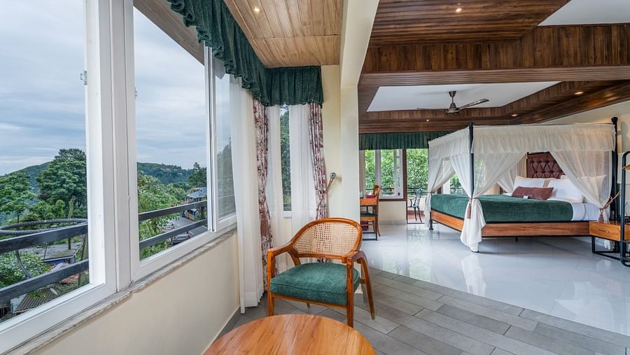 Large windows with nature views, comfortable seating, and a wooden table complement the spacious bed with side curtains and wooden furnishings