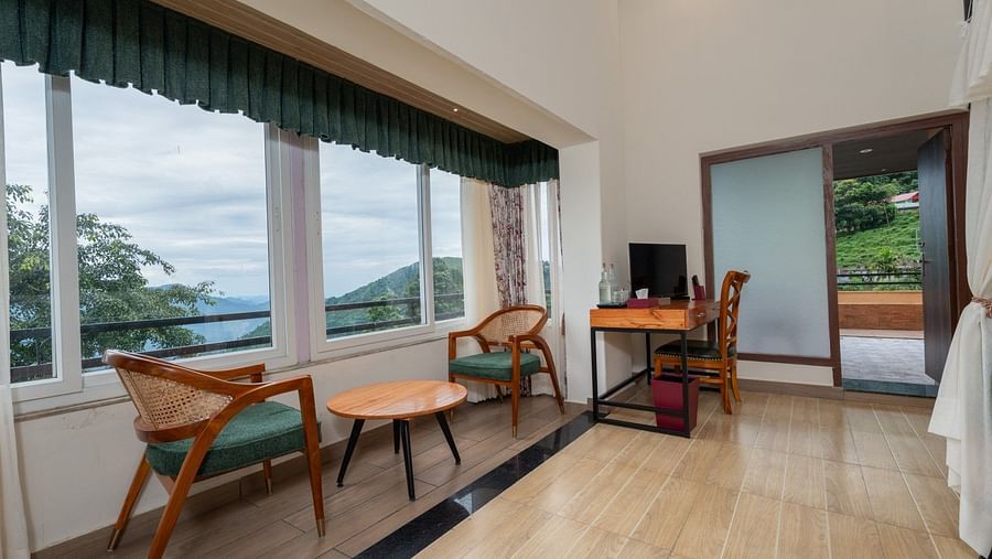 Large windows with nature views, comfortable seating, and a wooden table complement the spacious bed with side curtains and wooden furnishings