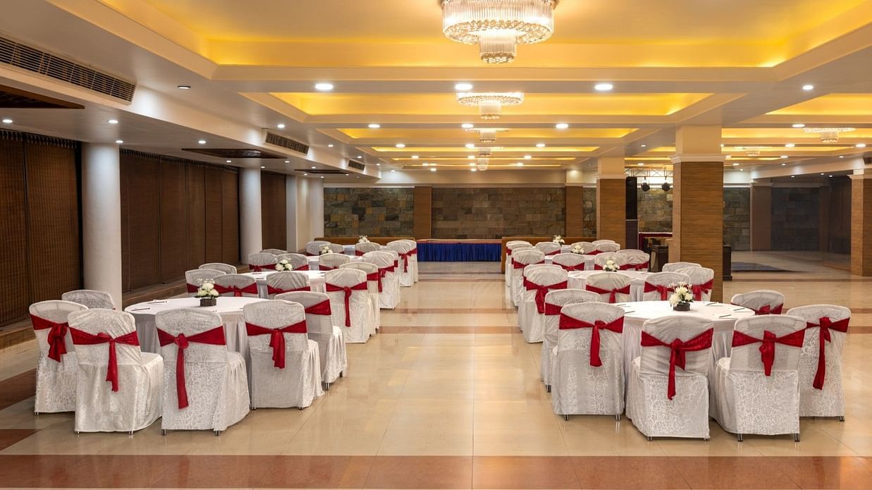 A spacious banquet hall arranged with round tables and white chairs adorned with red sashes. Warm recessed lighting and a central chandelier illuminate the room.  - Clarks Inn Express, Near Jammu Bari Brahmana Railway Station