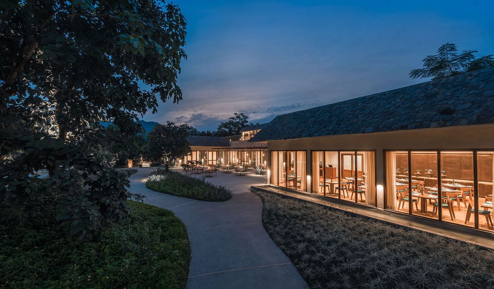 Exterior of a well-lit restaurant at Saraca  Resort & Spa Corbett with mountains in the backdrop during the blue hour