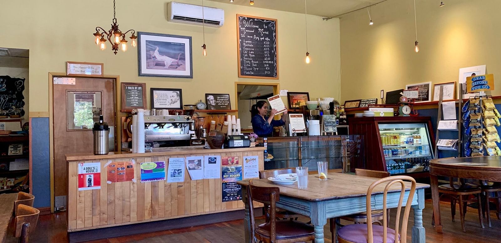 A room with tables and chairs features a counter with a coffee machine and various items on display, with framed pictures on the walls.