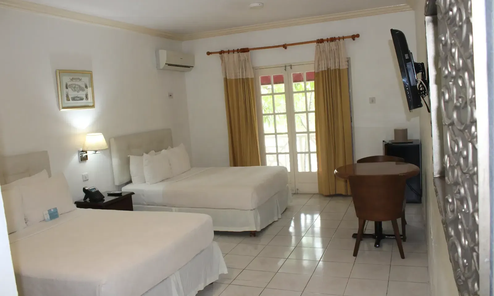 A clean hotel room includes a bed with white linens, a dark wooden dresser, and a window with light-colored curtains | Deluxe Double Room | Altamont Court, Jamaica