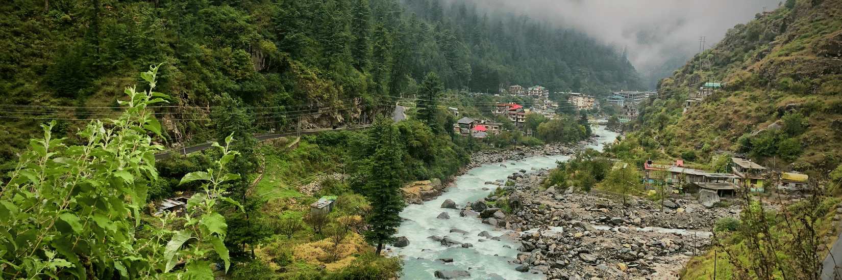 A panoramic view of the river valley in Kasauli, featuring a fast-flowing river cutting through a rocky landscape, surrounded by dense pine-covered hills. A small settlement is visible in the distance, with mist-covered mountains creating a scenic backdrop