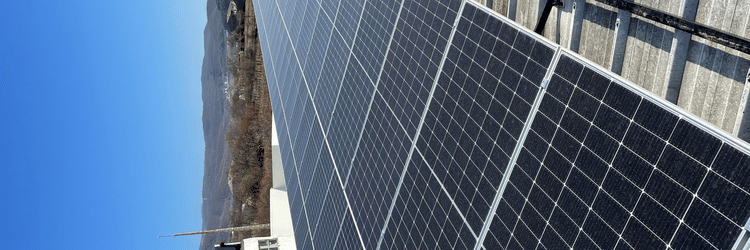 A long row of dark solar panels mounted on top of a building's roof, stretching toward a hilly landscape under a clear blue sky.