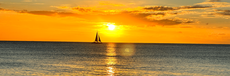 A sailboat drifting on calm ocean waters beneath a vibrant orange sunset sky visible from The Soco Hotel.