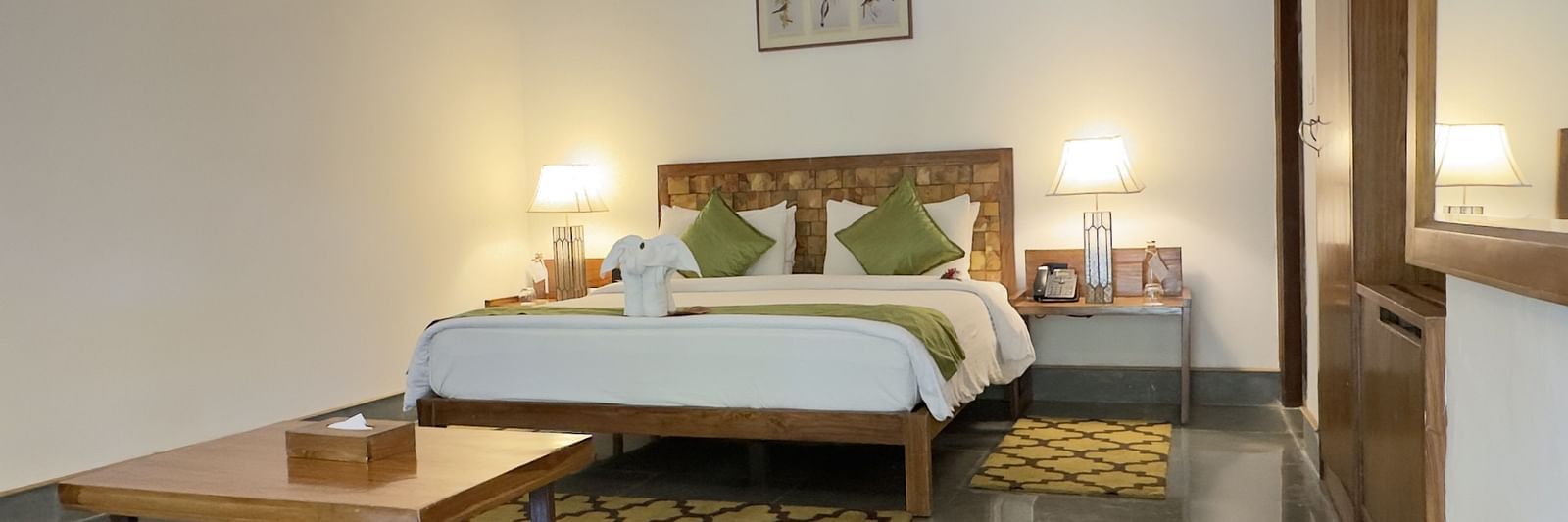 An image of a bedroom with double size bed, a centre table, bedside table on either side and lamps lit on top - Saraca Resort & Spa Corbett