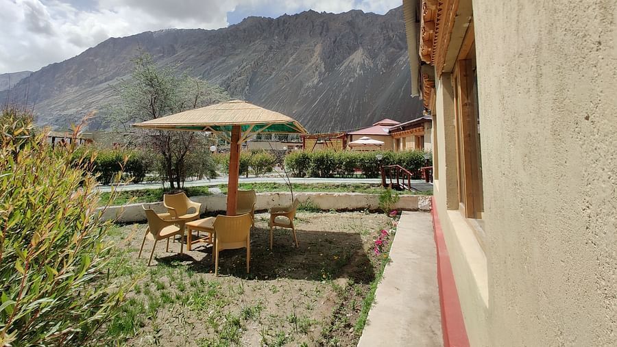 An outdoor seating area with a gazebo and chairs, adjacent to a building with a light-coloured exterior, with mountains visible in the background at White Sands Resort, Ladakh.