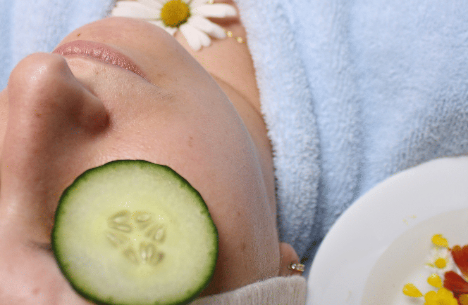 A person with a face cream and cucumber on their eye, next to a bowl of petals.