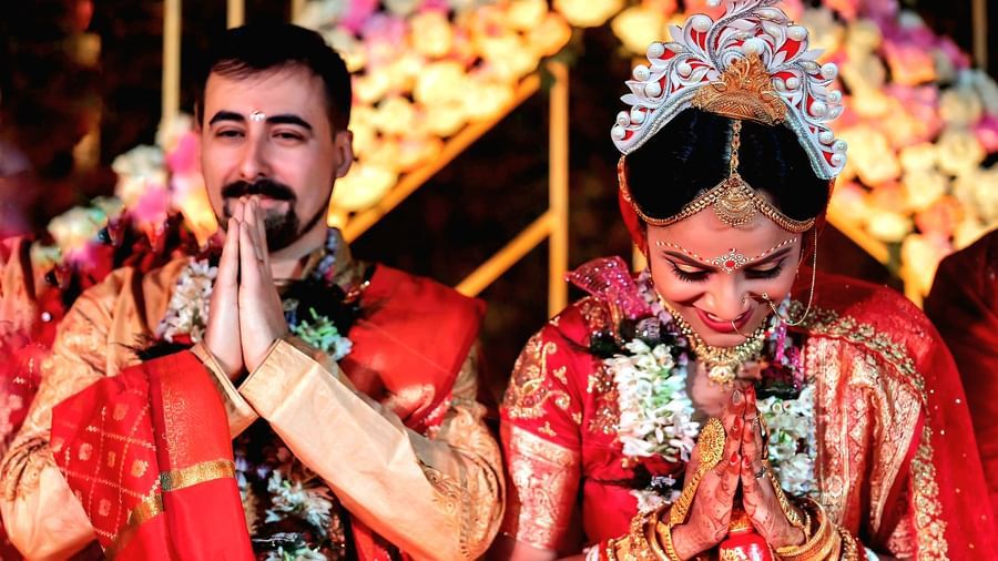 A couple in traditional Indian wedding attire performs a ritual with hands joined under a floral archway