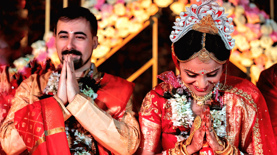 A bride and groom in traditional red wedding attire exchanging garlands under a decorated floral arch at Vedic Village Spa Resort with colorful flower decorations surrounding them.