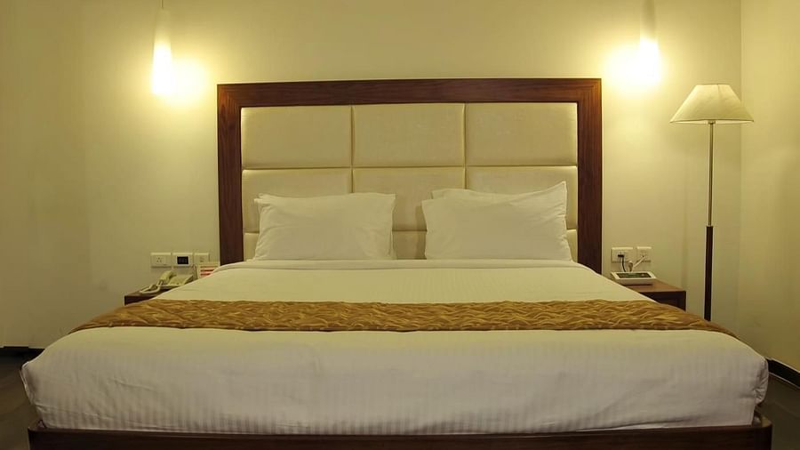A queen size bed with white linens and a gold runner, two hanging lights, a floor lamp, and a white rug on a wooden floor at Dhanalakshmi Srinivasan Hotels, Perambalur.