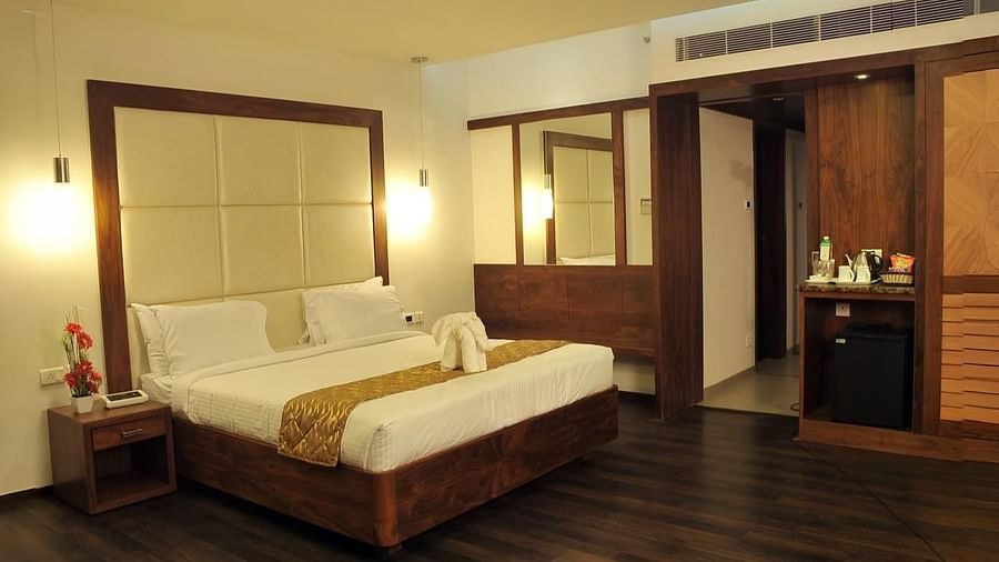 A room with a queen size bed, two side tables, wooden flooring, two mirrors, hanging lights, and a counter with bottles and appliances at Dhanalakshmi Srinivasan Hotels, Perambalur.