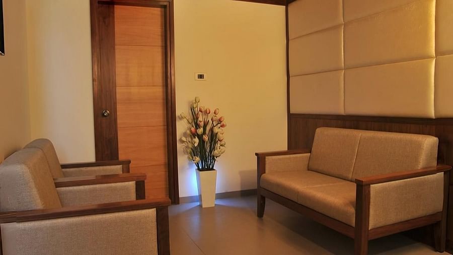 A lounge area with a wooden ceiling, two single-seater chairs, one two-seater sofa, a flower vase placed near a wooden door, and softly lit recessed lights on the ceiling.