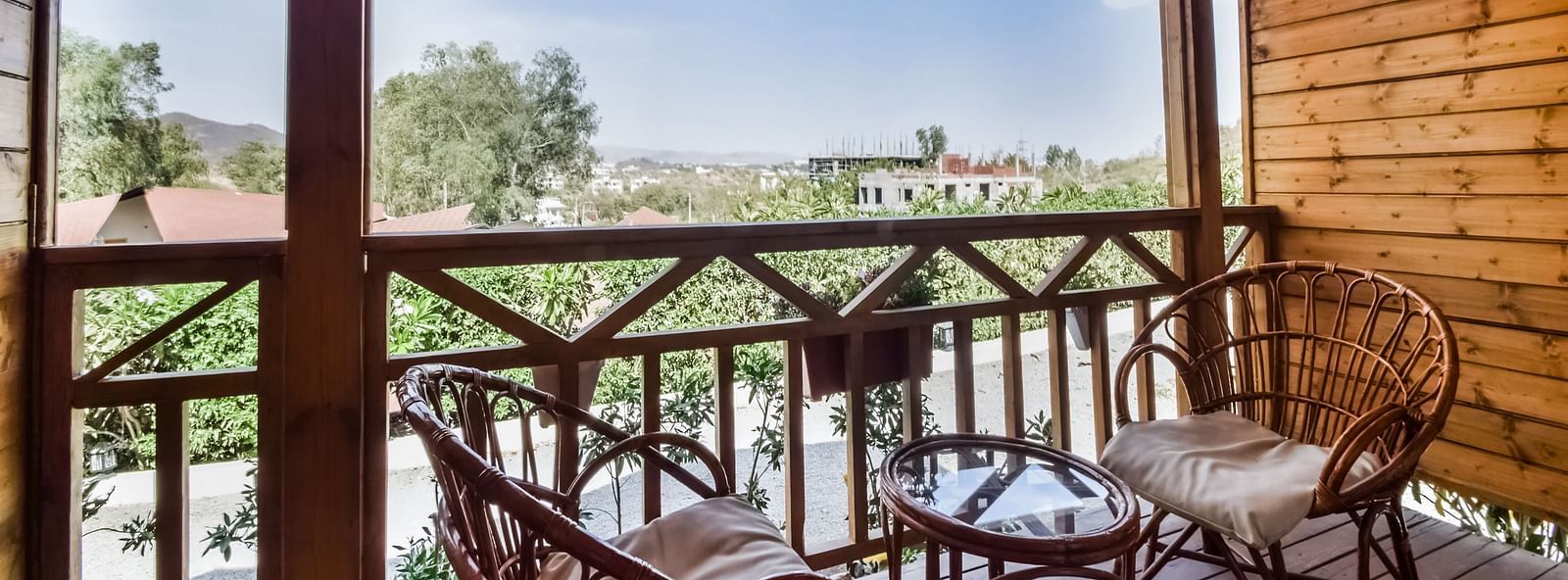 A wooden balcony with two wicker chairs and a small table overlooks a scenic view of trees .- Bamboo Saa Resort & Spa, Udaipur