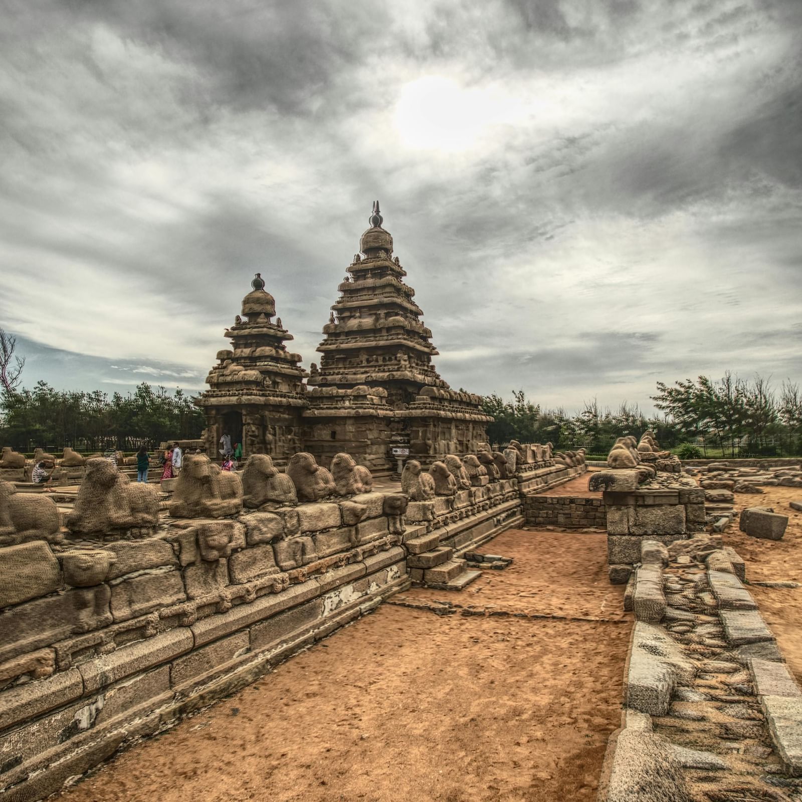 A wide-angle view of the Shore Temple complex, showing the ancient stone structures and a pathway leading towards them under a cloudy sky.