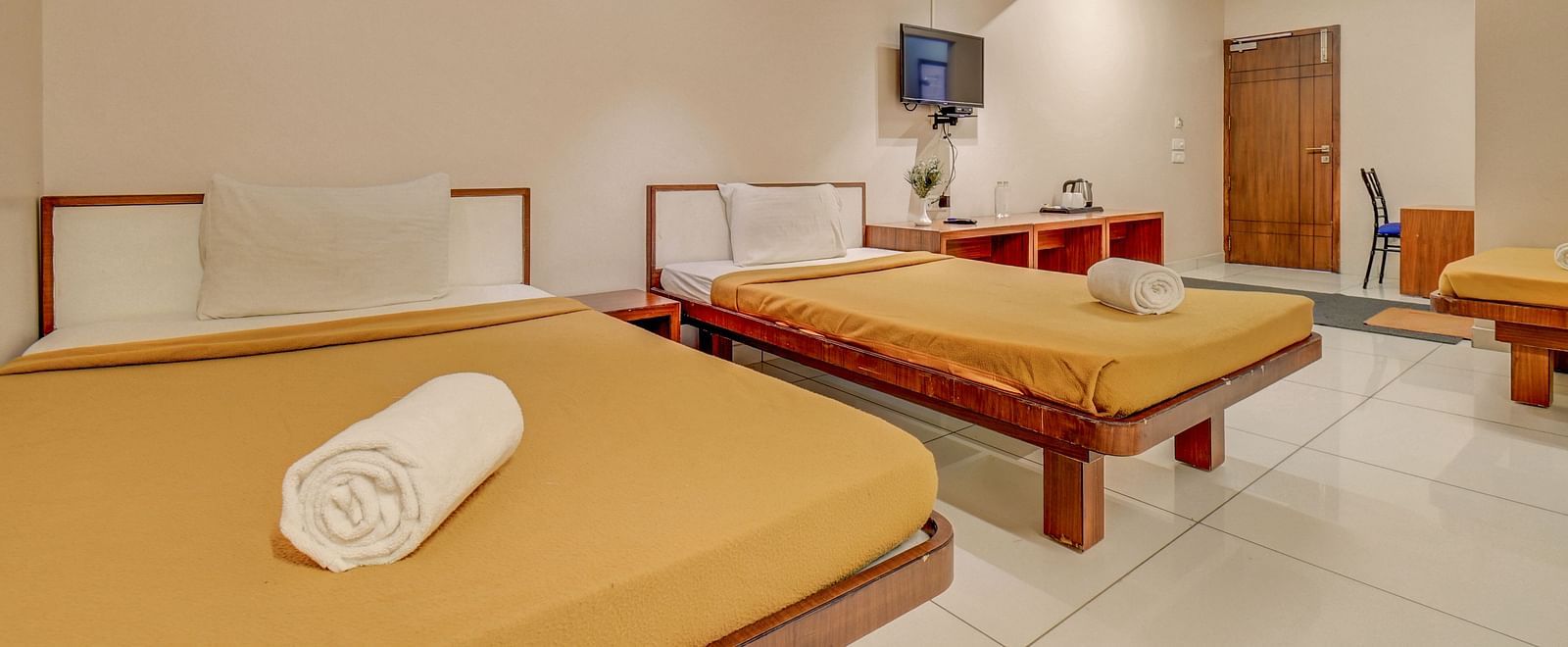 A spacious room at Hari Govinda’s JN Stay, Shakthinagar featuring neatly made single beds, a wall-mounted television, and a door at the furthest end