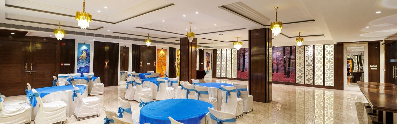 Banquet hall at Inde Reva Resorts Rishhikesh set up with round tables, white chairs, blue covers, and ceiling chandeliers.