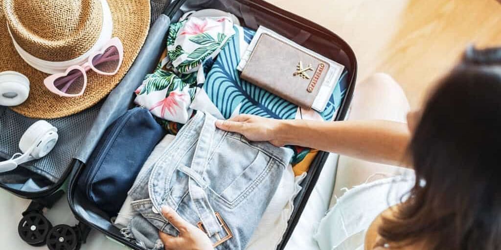 A person packing a suitcase with clothing, accessories like a sun hat, sunglasses, and a wallet, preparing for a trip.