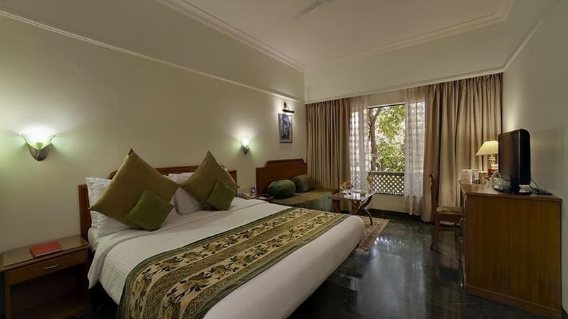 A comfortable hotel room at Renest Shraddha Inn features a bed with decorative pillows and a patterned runner, a seating area, a television on a stand, and large windows with curtains - Renest Shraddha Inn, Shirdi