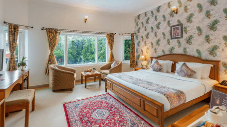 A bedroom at Amantra Shilpi Resort, Udaipur, with a king size bed, a work desk and chair, a red rug, and a window seating area.