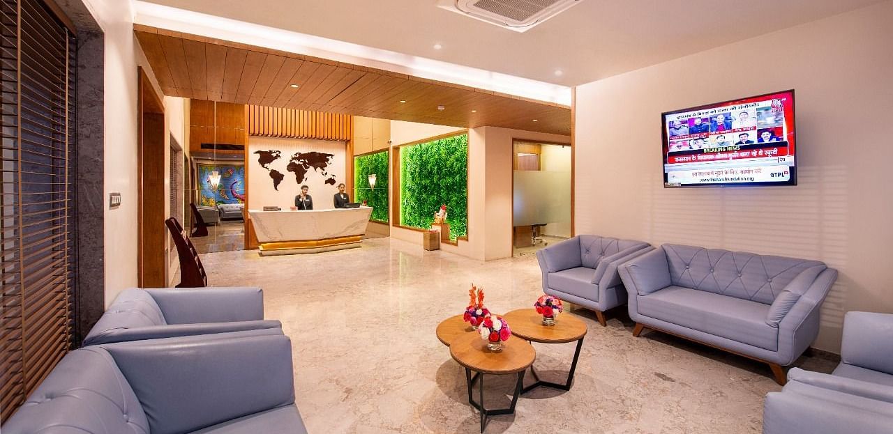 A comfortable hotel lobby lounge area with plush light blue sofas and armchairs arranged around small round tables with floral decorations, a television screen on the wall, and a view towards the reception desk with a world map backdrop and green wall feat