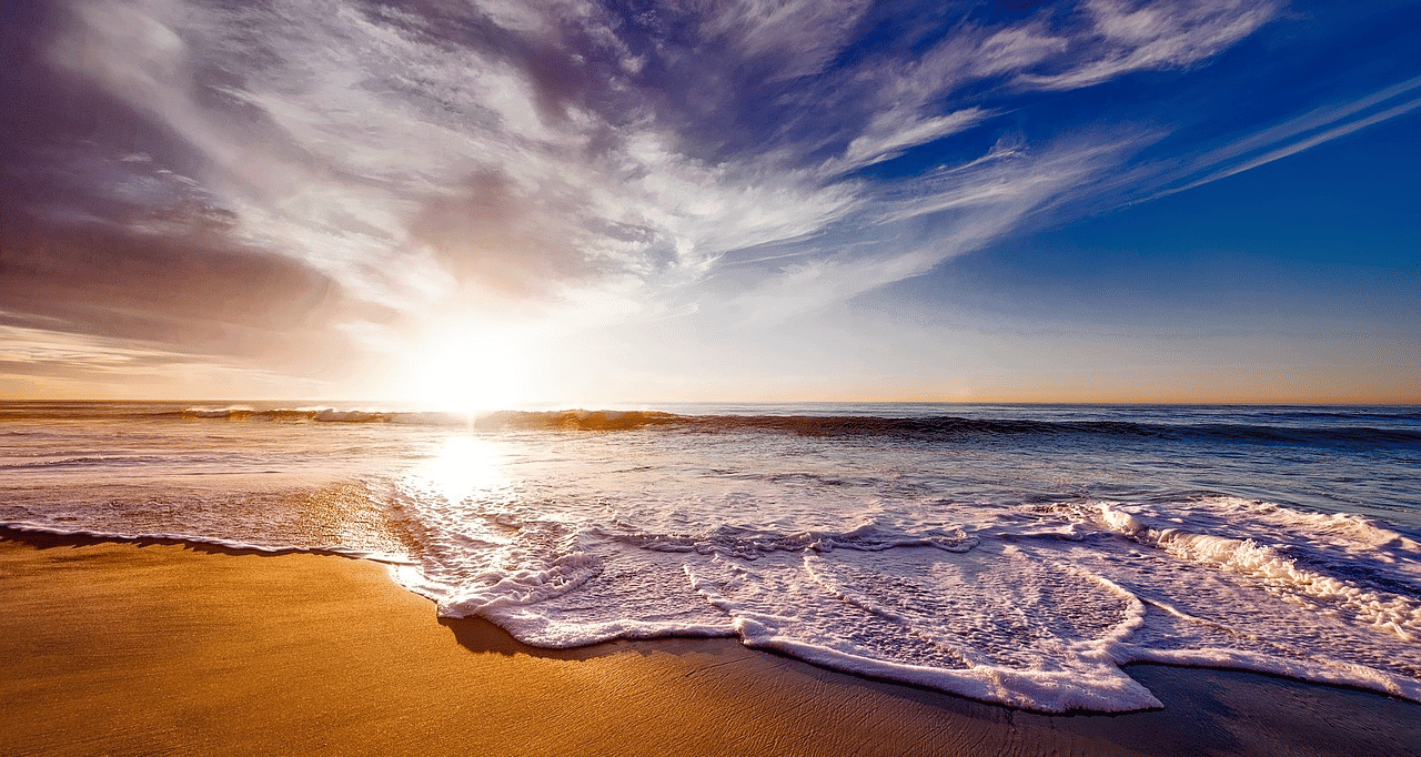 Waves crash onto a sandy beach under a dramatic sky with clouds as the sun sets over the ocean.