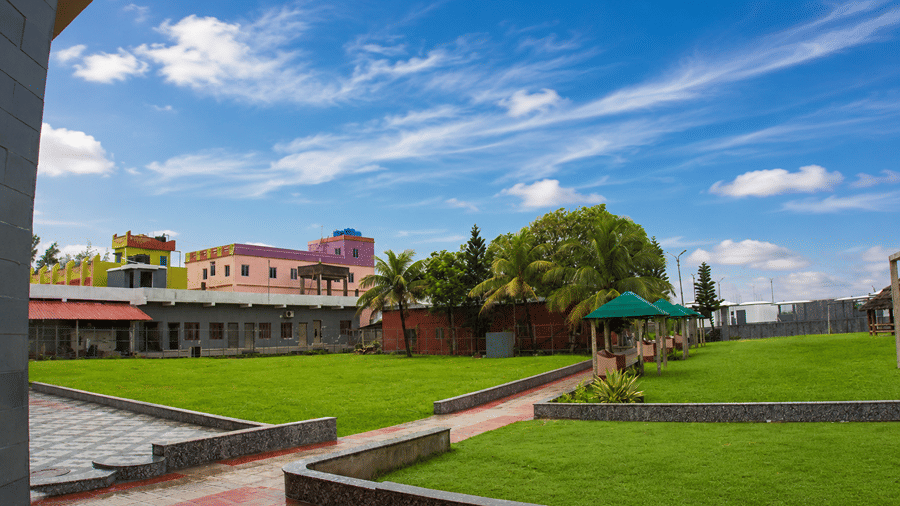 Manicured lawn at Pride Elite Mandarmani with green gazebos, a thatched hut, a stone pergola, and a paved pathway lined by coconut palms.