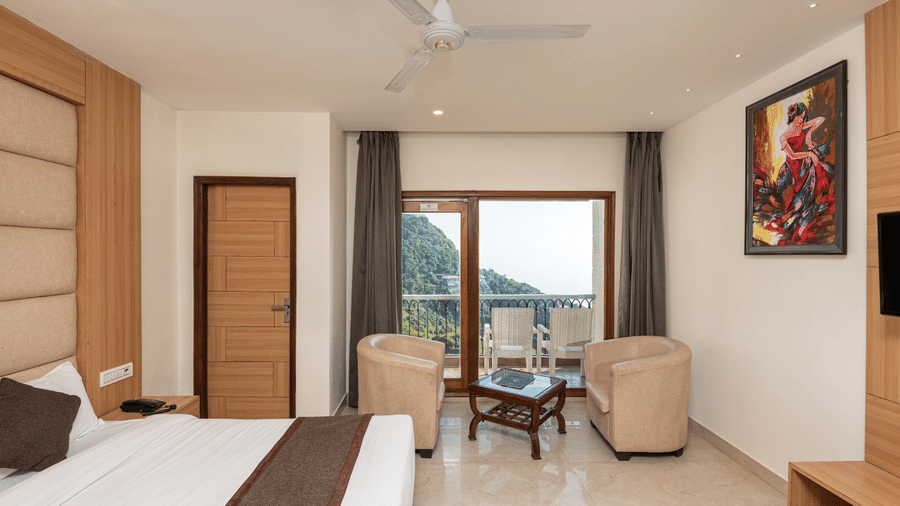 A modern hotel room at Shining Hills, Mussoorie with a double bed, ceiling fan, wall-mounted TV, and a glass window, offering view of hills.