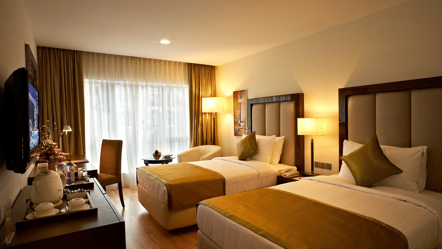 The Executive Twin bedroom featuring twin bed, bedside table with lamps, flat TV screen, a cabinet with tea and coffee maker and a work desk and chair at Adarsh Hamilton