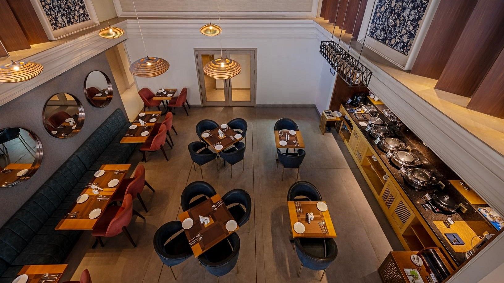 Overhead, the dining area glows warmly under yellow pendant lights, with red and blue chairs offering seating for two or four, a buffet on one side, and mirrored walls on the other.