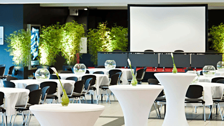 A small conference hall with a projector screen and white tables.