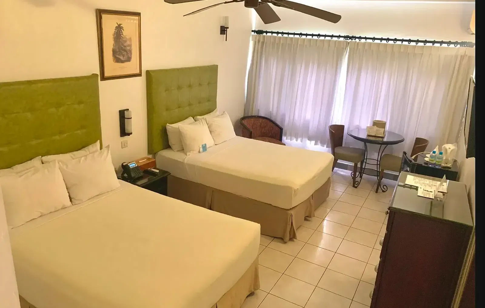 A hotel room showcases two beds with green headboards, a dresser, and a television, with a ceiling fan visible | Executive Double Room | Altamont Court, Jamaica