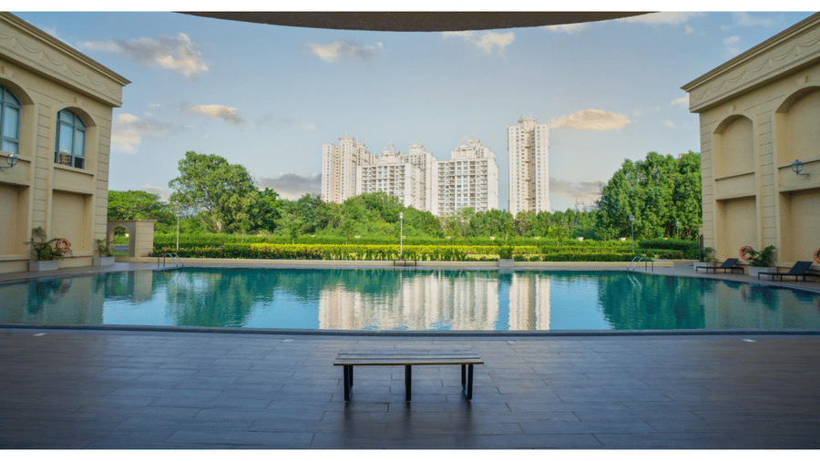 A large, tranquil outdoor swimming pool at a residential club house, reflecting the sky and surrounded by stone paving and distant tower blocks at Apartel by Aarin, Oragadam.