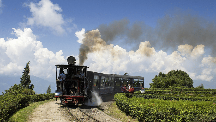 A view of a train travelling on a track with smoke coming out of the top and greenery surrounding the train, symbolising the Ooty toy train.